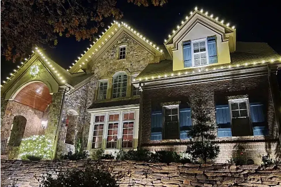 Stone and brick home with shutters, outlined gables and soft uplighting on the front wall.